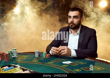 Ein Mann in einem Geschäftsanzug, der am Spieltisch sitzt. Männlicher Spieler. Leidenschaft, Karten, Chips, Alkohol, Würfel, Glücksspiel, Casino - es ist wie männliche Unterhaltung. Stockfoto