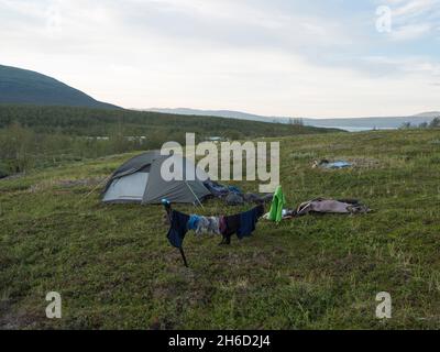 Grünes Trekkingzelt mit hängenden Kleidern in wunderschöner Natur mit Akka-Bergmassiv mit Schnee, Gletscher, Vuojatadno-Fluss und Birke auf Schwedisch Stockfoto