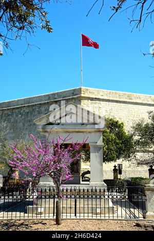 Maltesischer Flagge auf dem Dach St Johns Cavalier Gebäudes mit einer Gedenkstätte im Vordergrund in Hastings Gärten, Valletta, Malta, Europa. Stockfoto