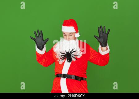 Eine Spielzeugspinne klettert durch den Bart eines erwachsenen kaukasischen Mannes, der als Weihnachtsmann gekleidet ist, während er erschreckt zuschaut. Der Hintergrund ist grün. Stockfoto