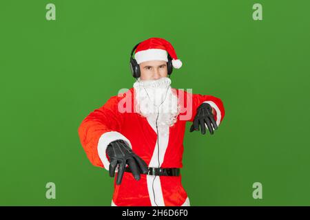 Ein erwachsener kaukasischer Mann, der als Weihnachtsmann mit Kopfhörern gekleidet ist, imitiert einen Discjockey, der vorgibt, die Schallplatten und die Lautstärkeregelung in der Luft zu bewegen. Stockfoto