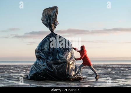 Eine Frau schiebt einen riesigen schwarzen Müllbeutel an das Ufer. Umweltverschmutzung durch Kunststoff. Wiederverwertbare Verpackung. Stockfoto