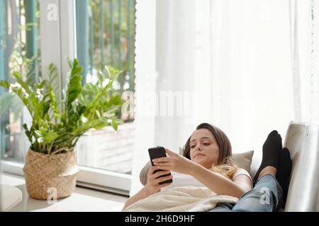 Hübsche junge Frau, die auf dem Sofa liegt, mit dem Gesicht neben den Füßen ihres Freundes und mit SMS-Nachrichten an Freunde Stockfoto