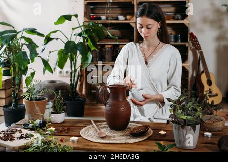 Wasser mit Kakao in den Krug gießen. Zubereitung von zeremoniellem Kakao in einem stimmungsvollen Café im Boho-Stil voller Pflanzen. Frau kocht gesundes Getränk aus Bio Stockfoto