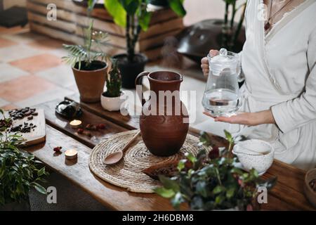 Wasser mit Kakao in den Krug gießen. Zubereitung von zeremoniellem Kakao in einem stimmungsvollen Café im Boho-Stil voller Pflanzen. Frau kocht gesundes Getränk aus Bio Stockfoto