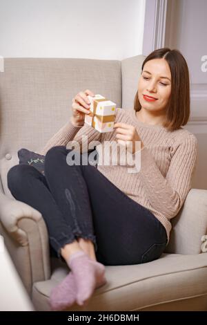 HÜBSCHE junge Frau, die in einem bequemen Sessel sitzt und eine Geschenkbox zu Hause hält. GHE rät, was drin ist Stockfoto