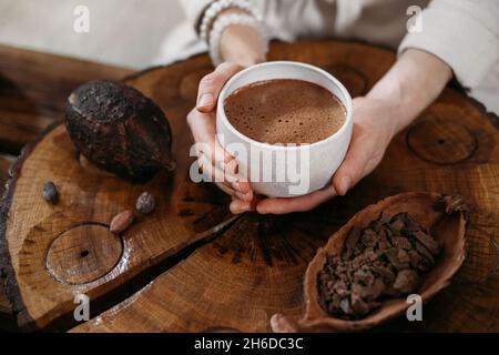 Heißer handgemachter zeremonieller Kakao in weißer Tasse. Frau Hände halten Handwerk Kakao, Draufsicht auf Holztisch. Bio gesundes Schokoladengetränk aus zubereitet Stockfoto