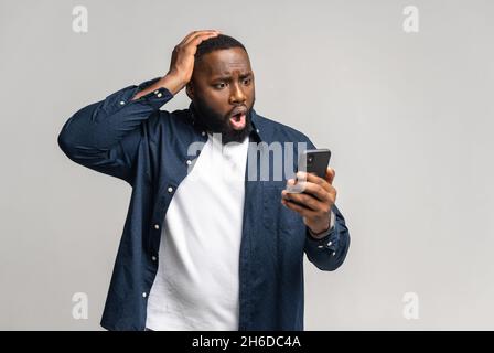 Schockiert afroamerikanischen Kerl Blick auf Smartphone-Bildschirm und halten Kopf verzweifelt über grauen Hintergrund. Multirassischer Mann erhielt unerwartete schlecht schockierte Nachrichten oder Benachrichtigungen. Copyspace, Werbung Stockfoto