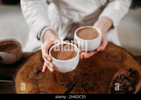 Heißer handgemachter zeremonieller Kakao in weißer Tasse. Frau Hände halten Handwerk Kakao, Draufsicht auf Holztisch. Bio gesundes Schokoladengetränk aus zubereitet Stockfoto