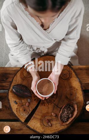 Heißer handgemachter zeremonieller Kakao in weißer Tasse. Frau Hände halten Handwerk Kakao, Draufsicht auf Holztisch. Bio gesundes Schokoladengetränk aus zubereitet Stockfoto