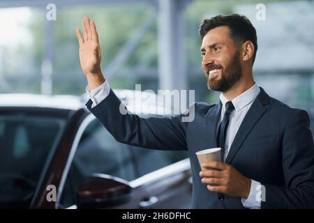 Bärtiger kaukasischer Mann in stilvollem Anzug lächelt und winkt jemandem zu, während er im modernen Autosalon steht. Geschäftsmann mit einer Tasse Kaffee in der Hand, der sich für ein neues Fahrzeug entschieden hat. Stockfoto