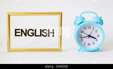 Das Wort ENGLISCH ist in einem goldenen Rahmen auf weißem Hintergrund neben dem Wecker geschrieben. Das Konzept der Bildung Stockfoto