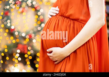 Bestes weihnachtsgeschenk. Schwangere in einem roten Kleid umarmt ihren Bauch vor dem Hintergrund des Weihnachtswohnzimmers in Unschärfe. Nahaufnahme kein Gesicht. Stockfoto