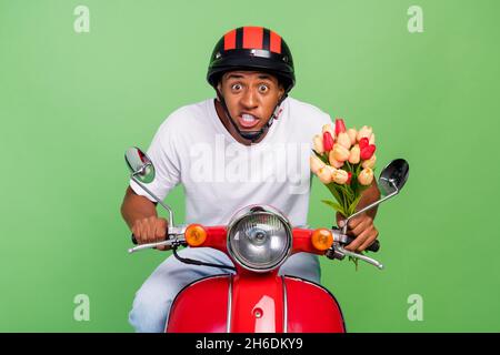 Foto von jungen afrikanischen Kerl schnelle Geschwindigkeit Eile Datum halten Blume Tulpen fahren Moped isoliert über grüne Farbe Hintergrund Stockfoto