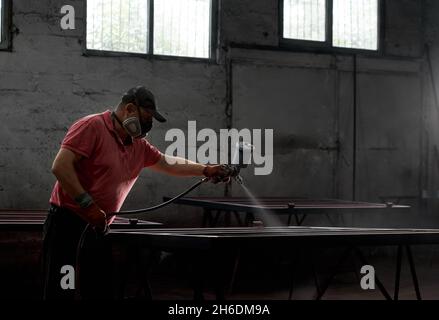 Kaukasischer Mann Malerei Metallkonstruktion mit Spritzpistole. Industriearbeiter trägt bei der Arbeit eine Schutzmaske und Handschuhe. Konzept von Service und Renovierung. Stockfoto