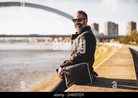Ein glücklicher Geschäftsmann sitzt draußen in der Stadt und genießt im Herbst die Sonne. Stockfoto