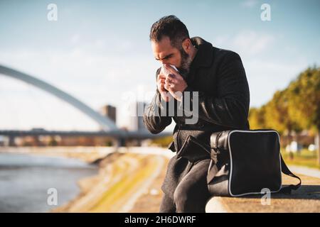 Der Geschäftsmann sitzt draußen in der Stadt. Er bläst seine Nase an einem kalten, sonnigen Tag im Herbst. Stockfoto