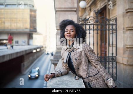 Schöne afroamerikanische junge Frau mit afro und großen Reifringen in einem stilvollen Mantel, lächelnd. Stadtleben Konzept, Street Portrait. Frühlings- oder Herbstzeit. Selektiver Fokus, Kopierbereich Stockfoto
