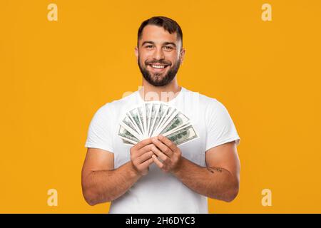 Happy Adult Attraktive kaukasischen Kerl in weißem T-Shirt halten eine Menge Dollar, isoliert auf gelbem Hintergrund Stockfoto