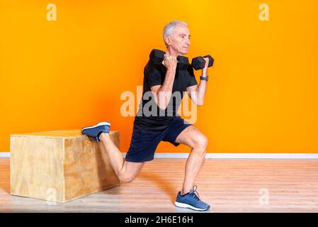 Fokussierter Senior fit Mann tun Gewichtheben Workout-Übung Stangen im Fitnessstudio isoliert auf orangefarbenem Hintergrund. Ältere Menschen Fitness-Training in Gebäuden. Stockfoto