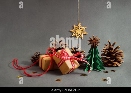 Origami Weihnachtsbaum und Papier Geschenkschachteln mit roten Bändern, kleinen Zapfen und goldenen Sternen auf grauem Hintergrund mit Strohstern, Weihnachtskomposition, Stockfoto