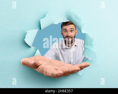 Glücklicher junger Kerl, der die Handfläche in zerrissenem blauem Papier durch ein Loch ausdehnt und ihm helfende Hand anbietet, um Unterstützung zu erhalten Stockfoto