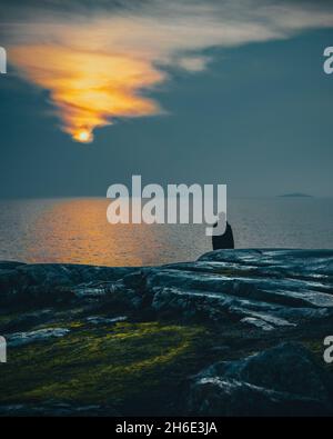 Mann, der allein auf dem Felsen steht, den Sonnenuntergang über dem Meer beobachtet und die friedliche Harmonie mit der Natur genießt. Sonne am Horizont. Silhouette der Person. Einsamkeit Stockfoto