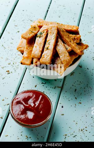 Nahaufnahme einer Glasschüssel mit Ketchup und einigen frittierten Streifen von Hühnchen in einer weißen Schüssel auf einem blauen Tisch Stockfoto