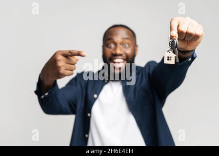 Selektiver Fokus auf die Schlüssel mit Schlüsselanhänger in Form von kleinen Haus in männlicher Hand. Lächelnder afroamerikanischer Mann hält Schlüssel von neuem Grundstück, zeigt Finger darauf, glücklicher Käufer von eigenem Anwesen, Kerl umgesiedelt Stockfoto