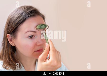 Junge kaukasische Frau tut Gesichtsmassage mit grünem Jade Roller auf neutralem Hintergrund Stockfoto