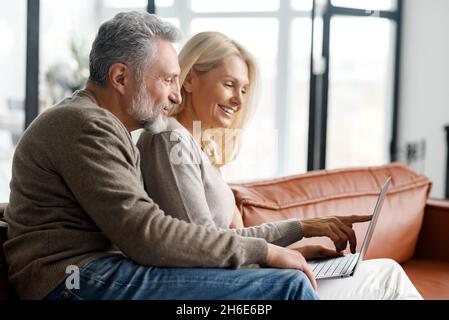 Das nette Paar mittleren Alters verbringt zuhause Freizeit online mit einem Laptop, die charmante Frau und der hübsche grauhaarige Mann sitzen auf der Couch, schauen sich Filme an oder kaufen gemeinsam online ein Stockfoto