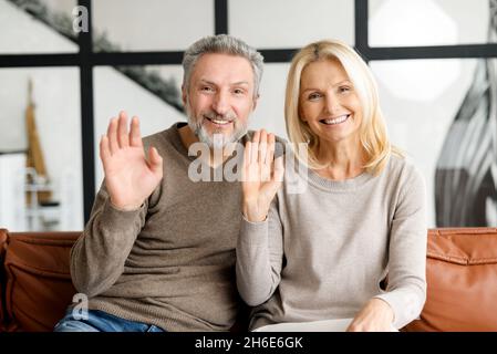 Video-Bildschirm mit fröhlichen Paar mittleren Alters, schaut auf die Kamera und winkt. Webcam-Ansicht einer lächelnden Frau und eines grauhaarigen Mannes, der auf der Couch sitzt und ein virtuelles Konferenzkonzept begrüßt Stockfoto