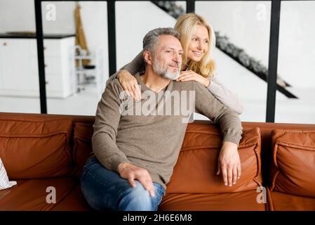 Porträt eines hübschen Paares mittleren Alters zu Hause, grauhaariger, hübscher Mann, der auf der bequemen Ledercouch sitzt, eine schöne blonde Frau, die sich von hinten umarmt. Reife Ehepartner verbringen Zeit miteinander Stockfoto