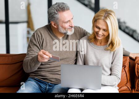 Fröhliches Paar mittleren Alters, das auf dem Laptop-Bildschirm zuschaut und die Kreditkartennummer überprüft, um Online-Transaktionen zu tätigen, lächelnde, reife Ehepartner kaufen online, überweisen Geld, Online-Banking Stockfoto