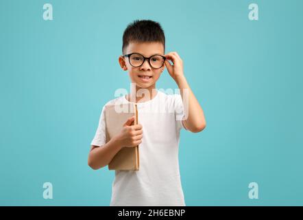 Portrait Of Little Asian Schoolboy In Brillen Mit Buch In Der Hand Stockfoto