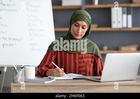 Muslimische Lehrerin im Kopftuch am Schreibtisch sitzend, schreibend, mit pc Stockfoto
