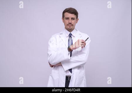 Porträt eines jungen kaukasischen Arztes in weißem medizinischen Gewand, der mit gekreuzten Armen steht. Isoliert auf Weiß. Kamera mit Kopierplatz betrachten Stockfoto