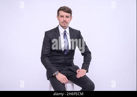 Porträt eines jungen kaukasischen, gutaussehenden Geschäftsmannes in schwarzem Anzug, der auf dem Stuhl sitzt. Isoliert auf weißem Hintergrund im Studio. Blick auf die Kamera Stockfoto