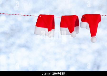 Drei rote kleine Weihnachtsmützen hängen an einem Seil auf einem verschwommenen, blau-grauen Hintergrund. Platz kopieren, Konzept Stockfoto
