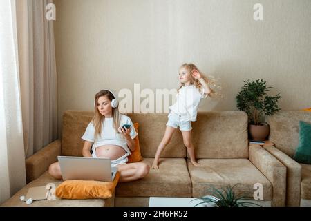 Beschäftigte Schwangere Mutter Freiberuflerin, die ferngesteuert mit Laptop Smartphone und Kopfhörer zu Hause auf der Couch arbeitet, während die Tochter tanzt und Spaß hat Stockfoto