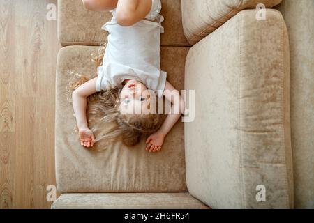 Porträt von kleinen blauäugigen 5 Jahre Mädchen Kind Kind mit lockigen blonden Haaren auf dem Sofa liegen. Kind Mädchen haben Spaß spielt liegen auf der Couch im Wohnzimmer an Stockfoto