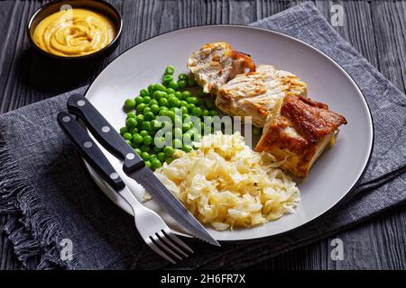 Gebratener Schweinelende und Sauerkraut auf einem Teller mit grünen Erbsen und Senf auf dunklem Holzboden, Draufsicht, Nahaufnahme Stockfoto