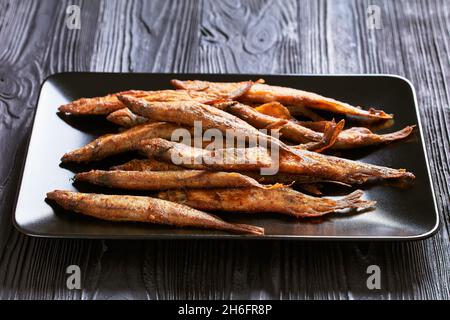 Knusprig gebratener goldbrauner Speer, Shishamo auf einer schwarzen rechteckigen Platte auf einem schwarzen strukturierten Holztisch Stockfoto