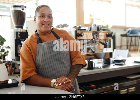 Porträt einer fröhlichen jungen erwachsenen afroamerikanischen Frau mit Schürze, die am Café-Tresen gegen Kaffeemaschinen steht und die Kamera anschaut Stockfoto