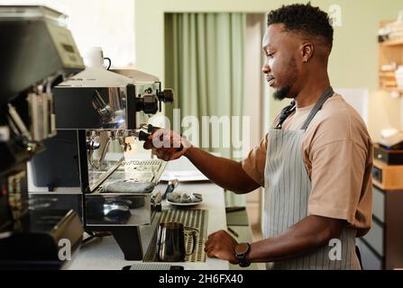 Horizontale Seitenansicht des hübschen jungen afroamerikanischen Mannes, der als Barista in einem kleinen Café arbeitet und Espresso mit einer modernen Kaffeemaschine zubereitet Stockfoto