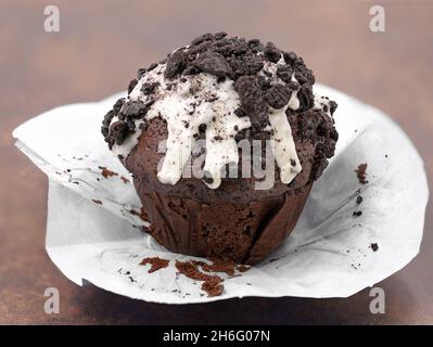 Schokoladenmuffin mit weißer Glasur und dunklen Keksbröseln auf braunem Hintergrund, ausgepackter Cupcake Stockfoto