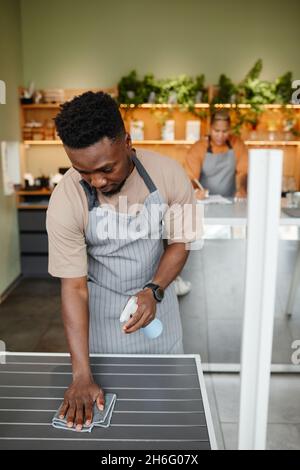Vertikale Aufnahme von modernen jungen Cafémitarbeitern, die mit der Reinigung von Möbeln und dem Erstellen von Notizen den Arbeitstag beginnen Stockfoto