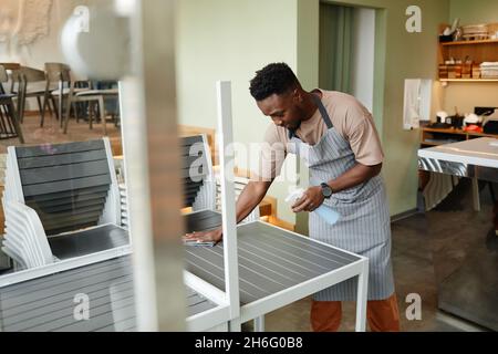 Junger afroamerikanischer Mann trägt Schürze ab Arbeitstag in einem modernen Café, der Tische für Kunden vorbereitet, die sie vor dem Öffnen reinigen Stockfoto