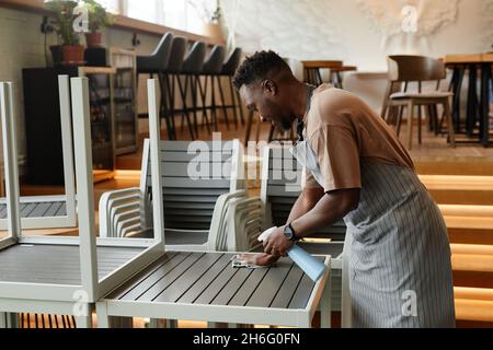 Junger afroamerikanischer Mann, der Schürze trägt, beginnt den Arbeitstag in einem kleinen Café, wo er Tische für die Kunden vorbereitet, die sie mit Waschmittel und Mikroib reinigen Stockfoto