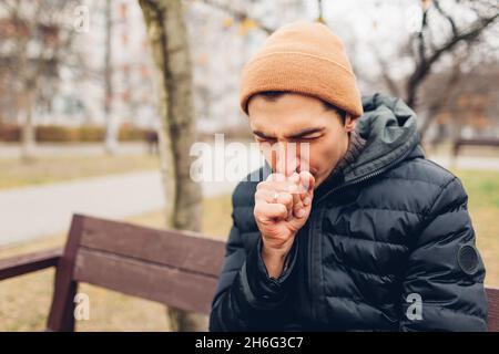 Junger kranker Mann hustet auf der Bank im Herbstpark, der den Mund bedeckt. Coronavirus covid-19 pandemische Grippe. Helferpflege im Herbst Stockfoto